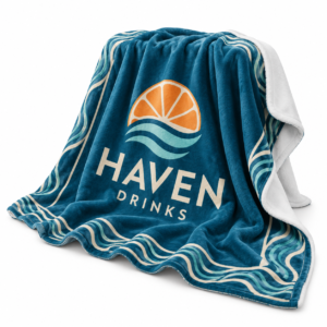 Draped teal fleece blanket with a colourful Haven Drinks logo, white reverse and soft folds on a white background